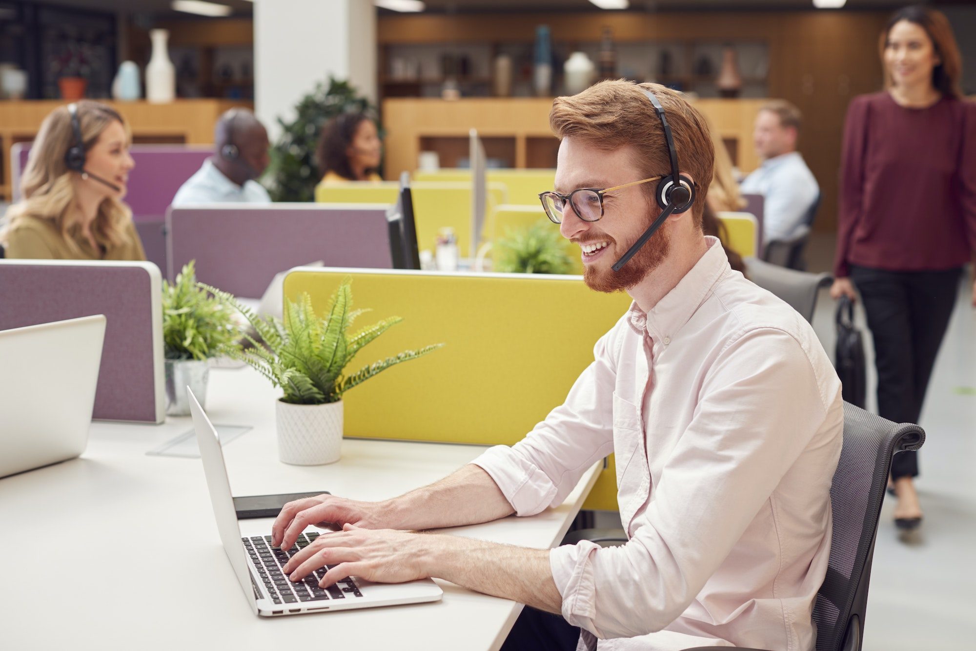 Businessman Wearing Headset Talking To Caller In Busy Customer Services Centre